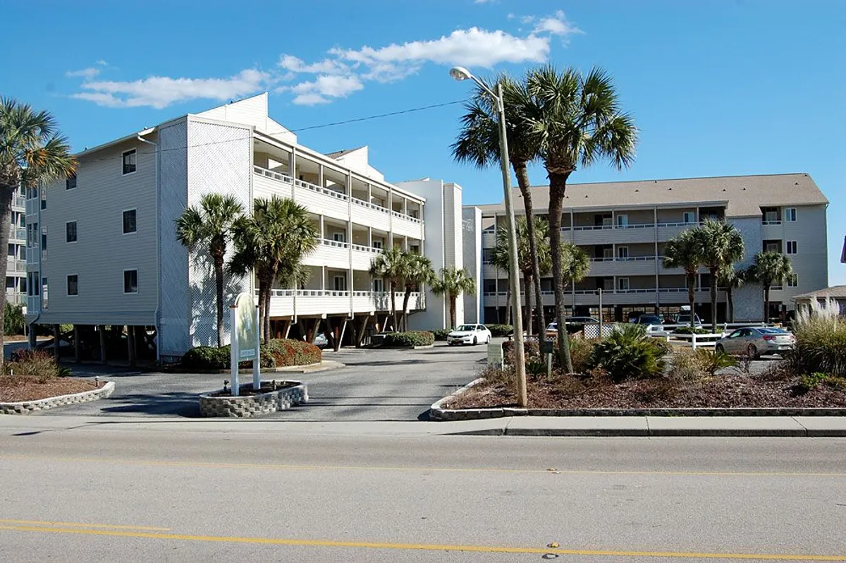 The image displays the exterior of the Pelican's Watch condominium complex, showcasing multi-story buildings surrounded by swaying palm trees and a neatly arranged parking area. The sunlit scene radiates a coastal, welcoming atmosphere, with hints of amenities just beyond the buildings and the promise of nearby beach access.