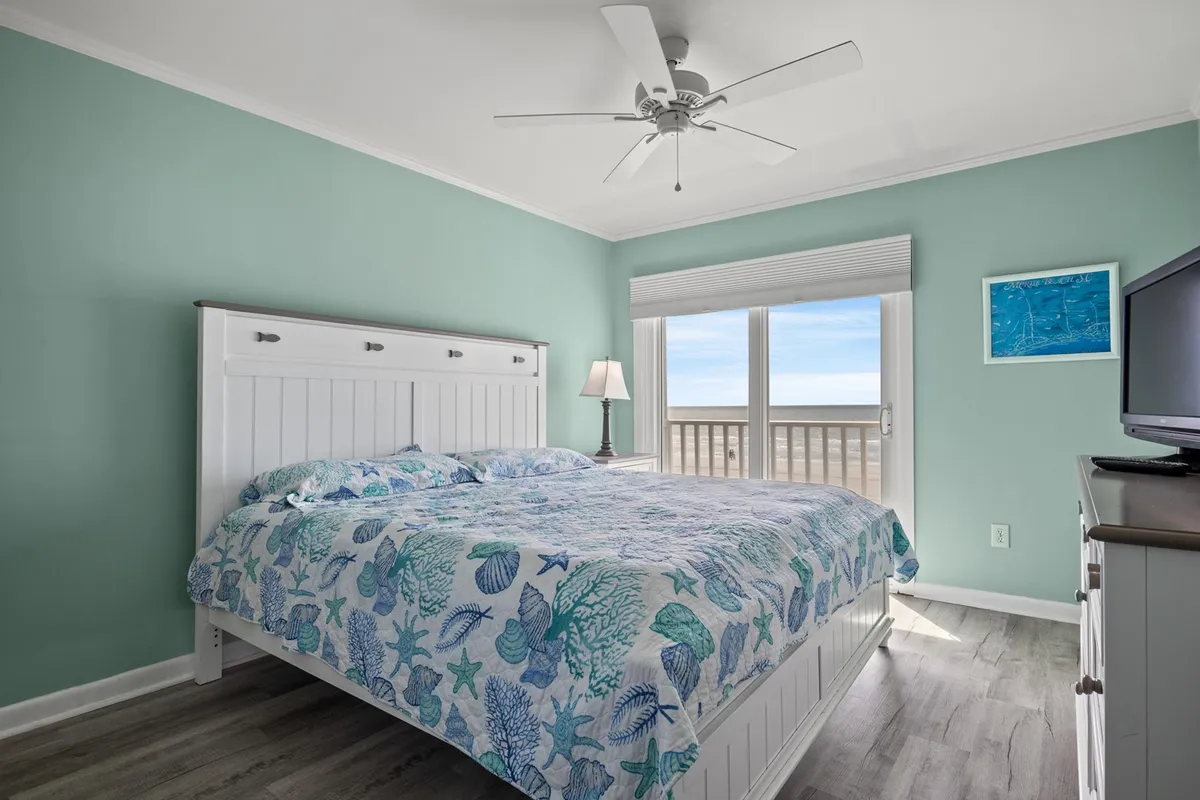 A serene bedroom featuring a bed adorned with a sea-themed quilt, large windows that let in plenty of natural light, and a bedside lamp creating a calm and restful ambiance. The soothing palette and ocean-inspired decor evoke a tranquil retreat, perfect for unwinding after a day of adventure.