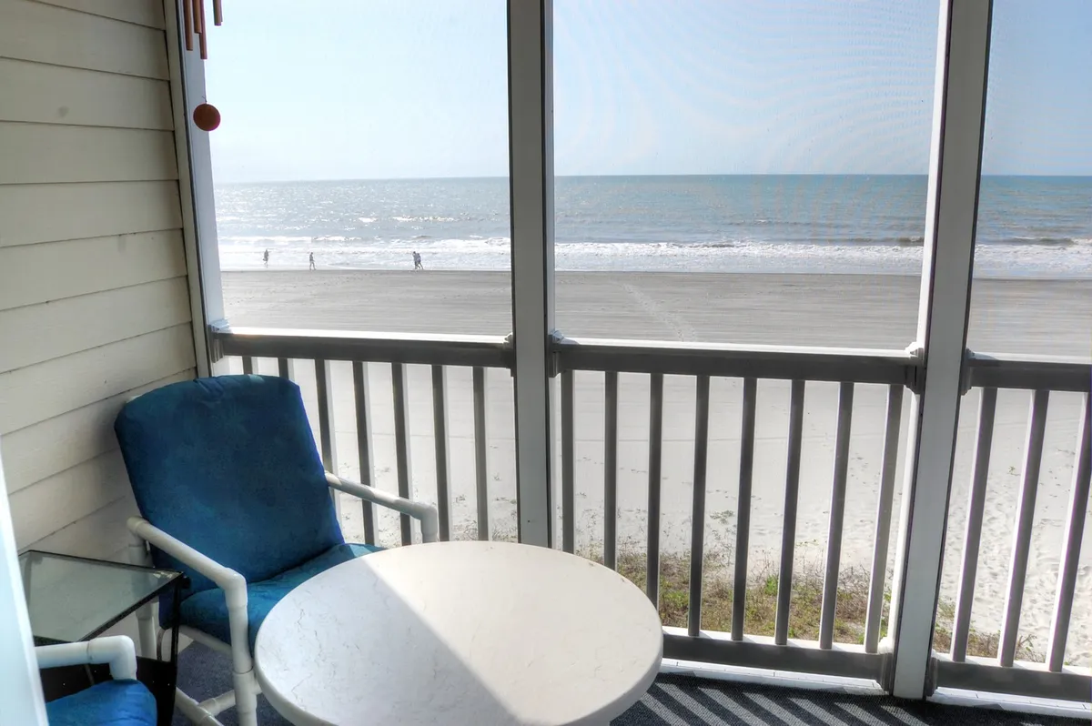A tranquil screened-in balcony with a seating area and table, offering a sweeping view of the beach and ocean beyond. Sunlight pours into the space, and the inviting ambiance encourages quiet mornings or contemplative evenings while gazing at the water.