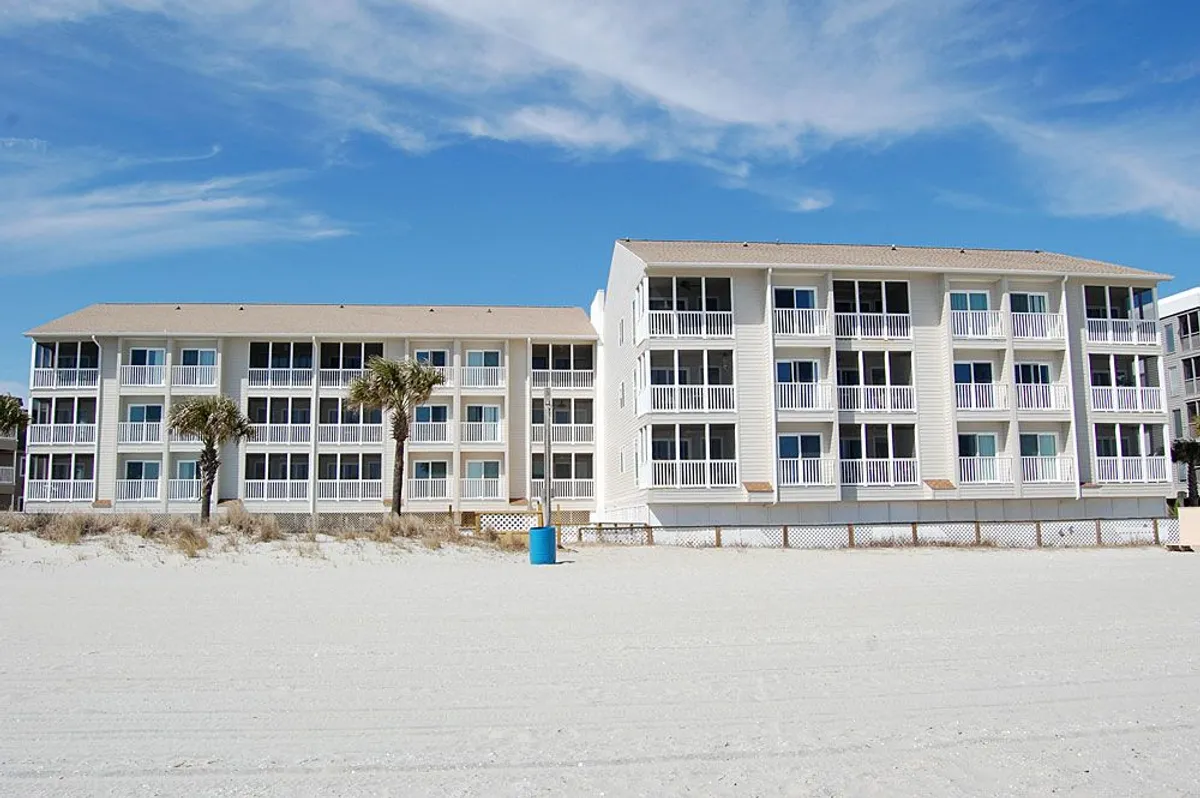 An exterior shot showing a beachfront view with palm trees and balconies overlooking the ocean. The scene captures guests relaxing on the beach and socializing on balconies, highlighting the proximity to the sand and the outdoor communal atmosphere.