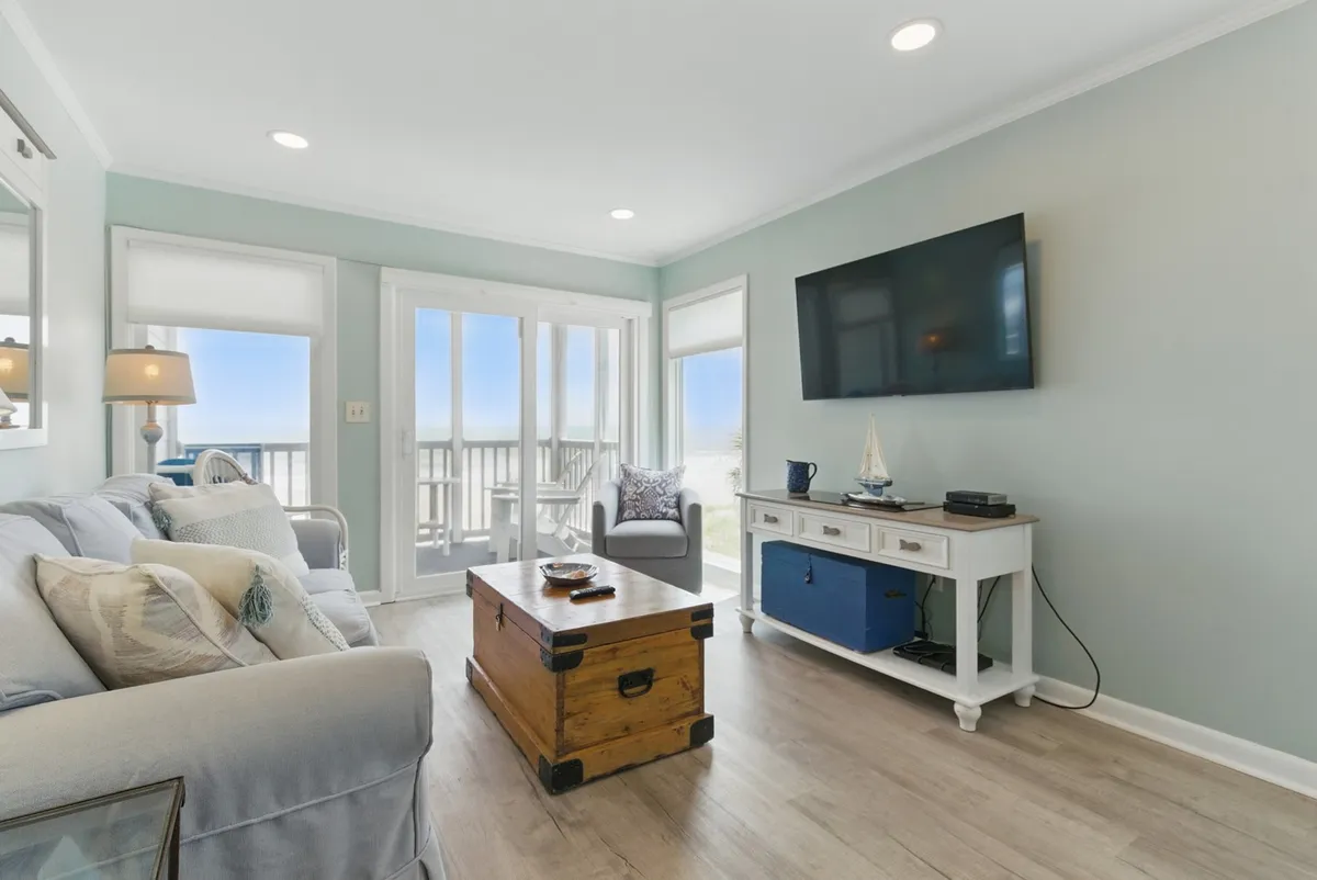 The image captures a comfortable living room with a sofa adorned with decorative pillows, a wooden coffee table, television, and large glass doors that open onto an outdoor deck. Ample natural light pours in, highlighting the open feel of the space. The setup encourages family gatherings, relaxation, and game nights, with the outdoors just a step away.