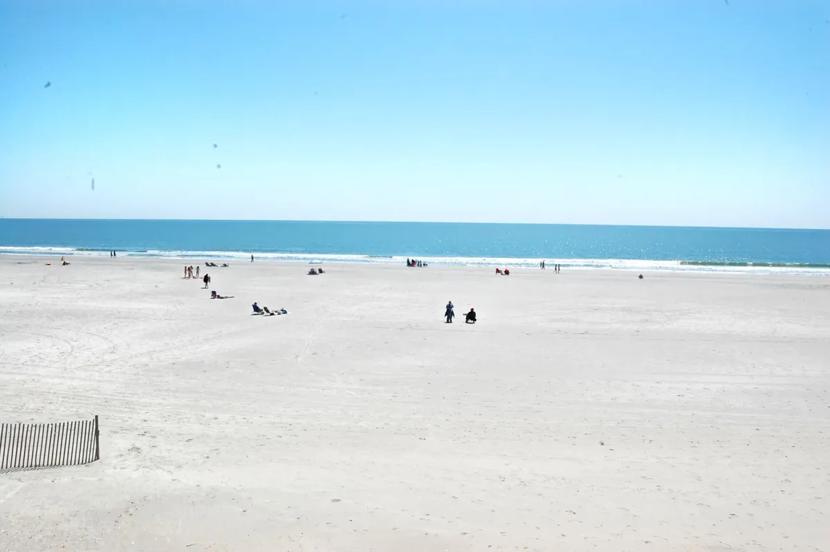 A wide view of the vast beach and calm ocean, with distant figures walking or sunbathing, underlines the broad, open coastal landscape. The scene is serene and spacious, evoking a sense of relaxation and possibility.