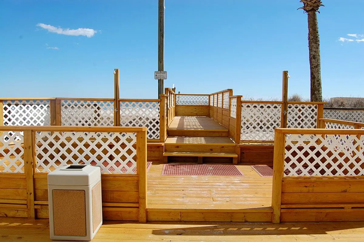 A wooden deck with lattice fencing and a palm tree frames the view, with a signpost suggesting welcoming community spaces. The deck appears to overlook the ocean, offering a spot for relaxation or watching the sunset.
