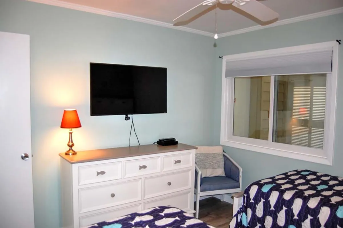 A cozy living room scene featuring a television, dresser, lamp, and a window streaming in natural light. The setup invites relaxation, with a comfortable ambiance suited for reading, movie nights, or unwinding after a day of exploring Myrtle Beach.